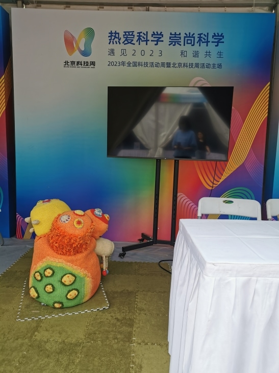 写有“北京科技周”字样的背景板、电视屏幕、桌椅，以及放在草绿色地毯上的毛绒玩具装置。