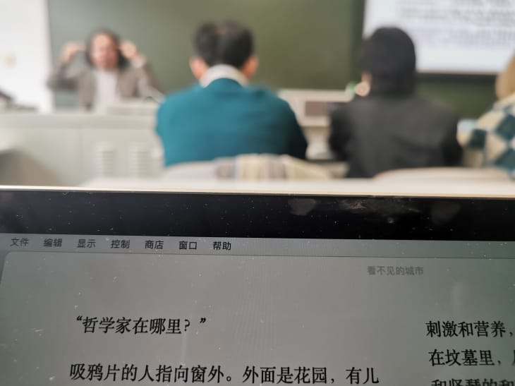 电脑屏幕上的电子书，第一行“哲学家在哪里？”。远处是讲台上老师的模糊影像。