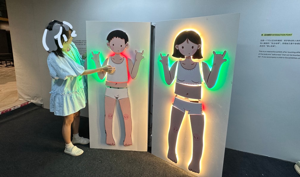 Full image of the installation. Images of two children on a textbook-shaped display panel. When touched with the hand, parts of the bodies light up with different colours surrounding their borders.