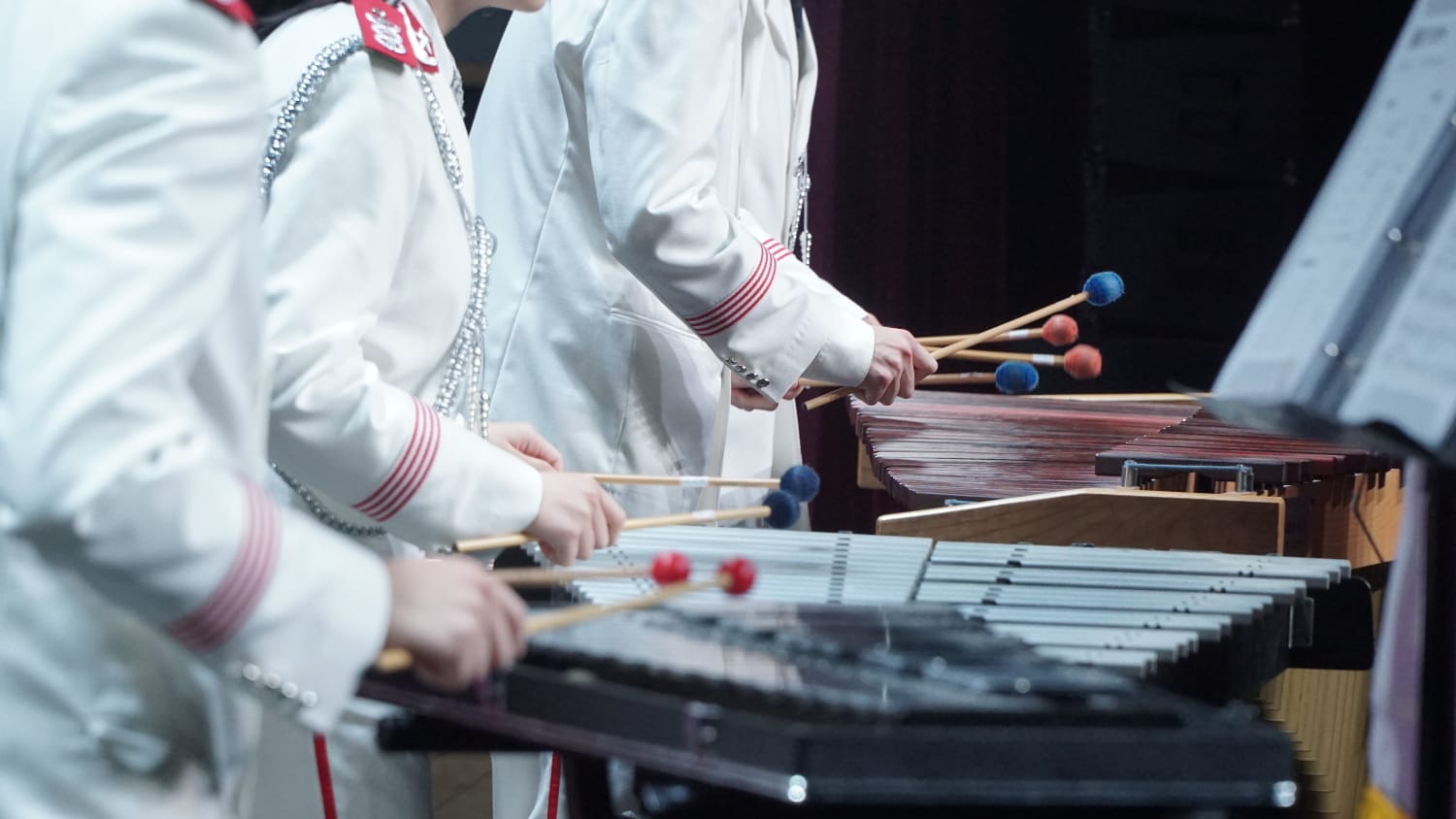 Low-angle side view photograph. On a stage against a black background, band members in white uniforms holding mallets and playing.