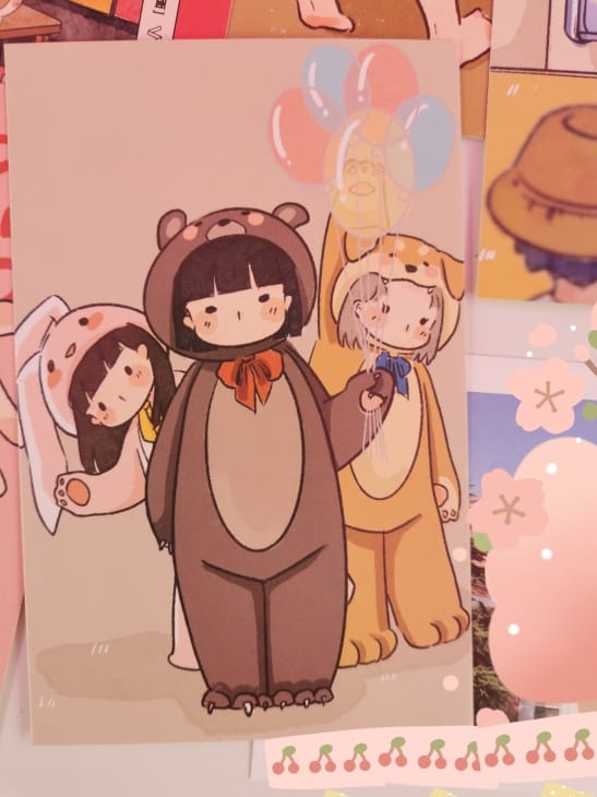 明信片一面的绘画：身着小动物服饰的三位女孩，其中一名手里拿着彩色气球。