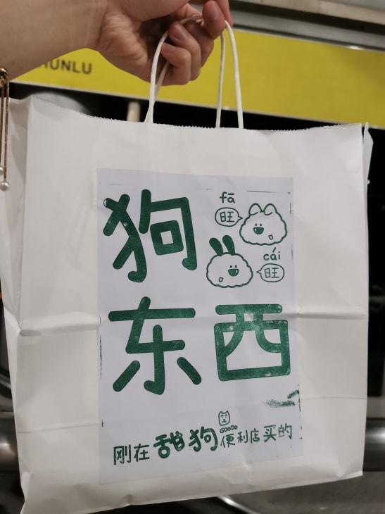 手提纸袋，写着大字“狗东西”，下面是“刚在甜狗便利店买的”。旁边绘制的卡通小狗说出“旺旺”，但是拼音是“发财”。